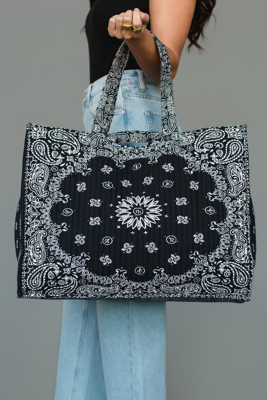 A person wearing a black top and light blue jeans holds the Panache Apparel Collection Black Bandana Tote, a large rectangular bag featuring a black and white paisley bandana print and two handles, against a gray background.