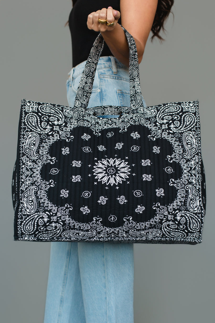 A person wearing a black top and light blue jeans holds the Panache Apparel Collection Black Bandana Tote, a large rectangular bag featuring a black and white paisley bandana print and two handles, against a gray background.