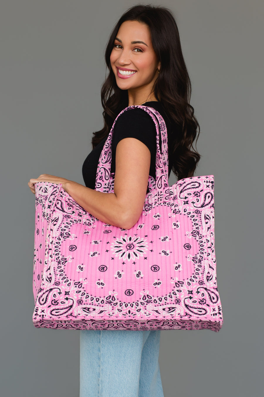 A woman wearing a black shirt and jeans smiles while holding the Panache Apparel Collection Pink Bandana Tote, featuring a large size and paisley bandana pattern, against a plain gray background.