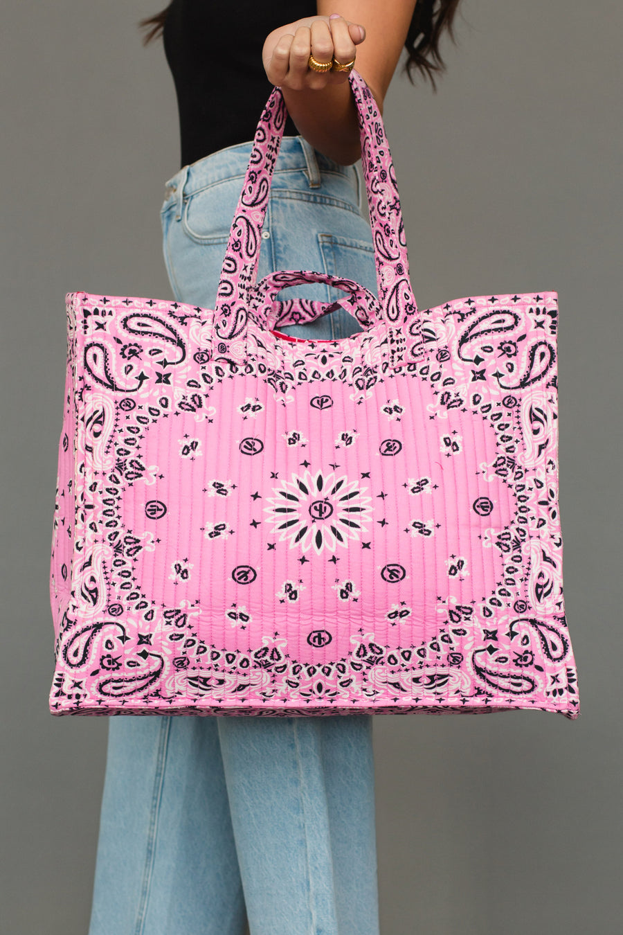 A person dressed in jeans and a black top holds the Pink Bandana Tote from Panache Apparel Collection, featuring a bold black and white paisley bandana pattern, set against a plain gray background.