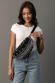 A woman wearing a white t-shirt and blue jeans smiles while holding the Panache Apparel Collection Black Bandana Crossbody over her shoulder, standing against a plain gray background.