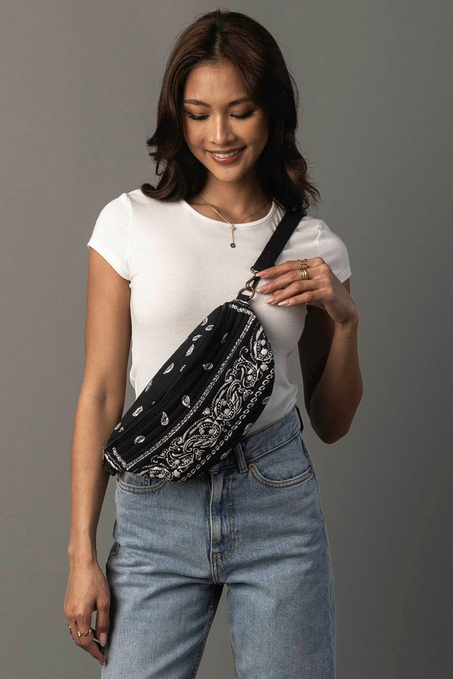 A woman wearing a white t-shirt and blue jeans smiles while holding the Panache Apparel Collection Black Bandana Crossbody over her shoulder, standing against a plain gray background.