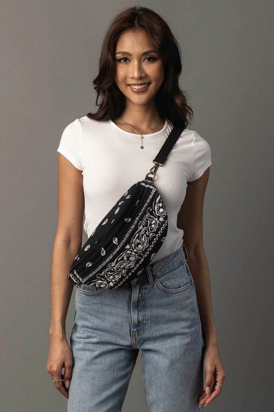 A woman in a white t-shirt and jeans smiles at the camera, wearing the Panache Apparel Collection Black Bandana Crossbody. She has dark, wavy hair, and stands against a plain gray background.