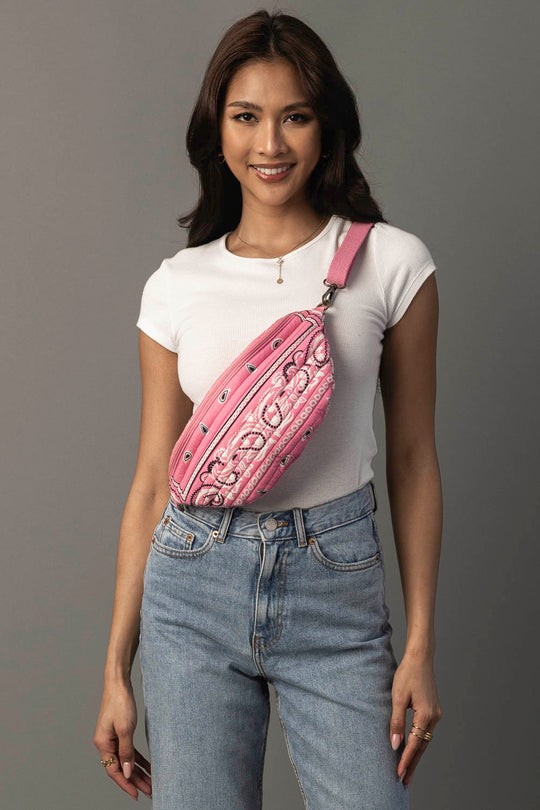 A woman wearing a white t-shirt and blue jeans smiles while posing with the Pink Bandana Crossbody from Panache Apparel Collection against a gray background.
