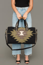 A person in light blue jeans and a black top holds the Panache Apparel Collection Black, Olive, Gray & Cream Western Duffel, featuring a geometric tribal pattern and decorative straps, standing against a plain background.