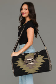 A woman with long dark hair, dressed in a black top and light blue jeans, stands sideways and smiles while carrying the Panache Apparel Collection Black, Olive, Gray & Cream Western Duffel over her shoulder.