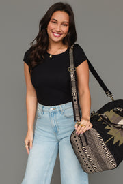 A smiling woman with long brown hair in a black top and light jeans holds the Panache Apparel Collection Black, Olive, Gray & Cream Western Duffel, featuring a geometric strap, while standing against a plain gray background.