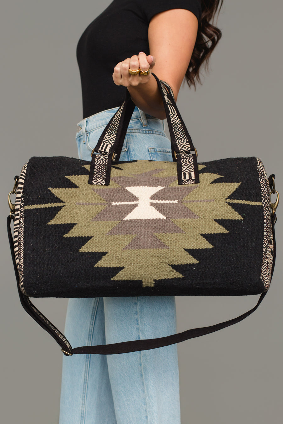 A person wearing a black top and light blue jeans holds the Panache Apparel Collection Black, Olive, Gray & Cream Western Duffel against a plain gray background.