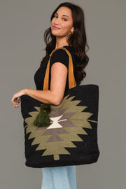 A woman with long dark hair, dressed in a black t-shirt and light blue jeans, smiles against a plain gray background as she holds the Panache Apparel Collection’s Black, Olive, Gray & Cream Western Tote, a spacious bag featuring a geometric pattern with tan straps and a green tassel.