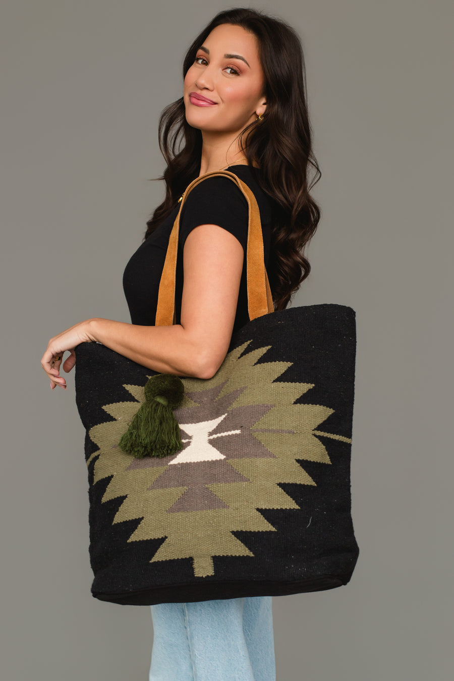 A woman with long dark hair, dressed in a black t-shirt and light blue jeans, smiles against a plain gray background as she holds the Panache Apparel Collection’s Black, Olive, Gray & Cream Western Tote, a spacious bag featuring a geometric pattern with tan straps and a green tassel.
