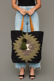 A person wearing a black top, light blue jeans, and black heels holds the Panache Apparel Collection’s Black, Olive, Gray & Cream Western Tote, featuring leather straps and a green tassel. Only the lower half of their body is visible.