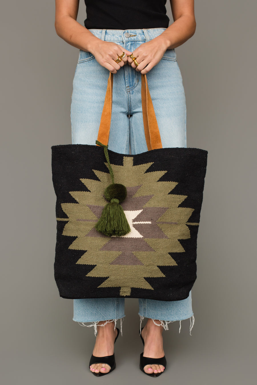 A person wearing a black top, light blue jeans, and black heels holds the Panache Apparel Collection’s Black, Olive, Gray & Cream Western Tote, featuring leather straps and a green tassel. Only the lower half of their body is visible.
