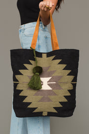 A person in light blue jeans and a black top holds the Panache Apparel Collection’s Black, Olive, Gray & Cream Western Tote, which features geometric patterns, tan handles, and green tassels.