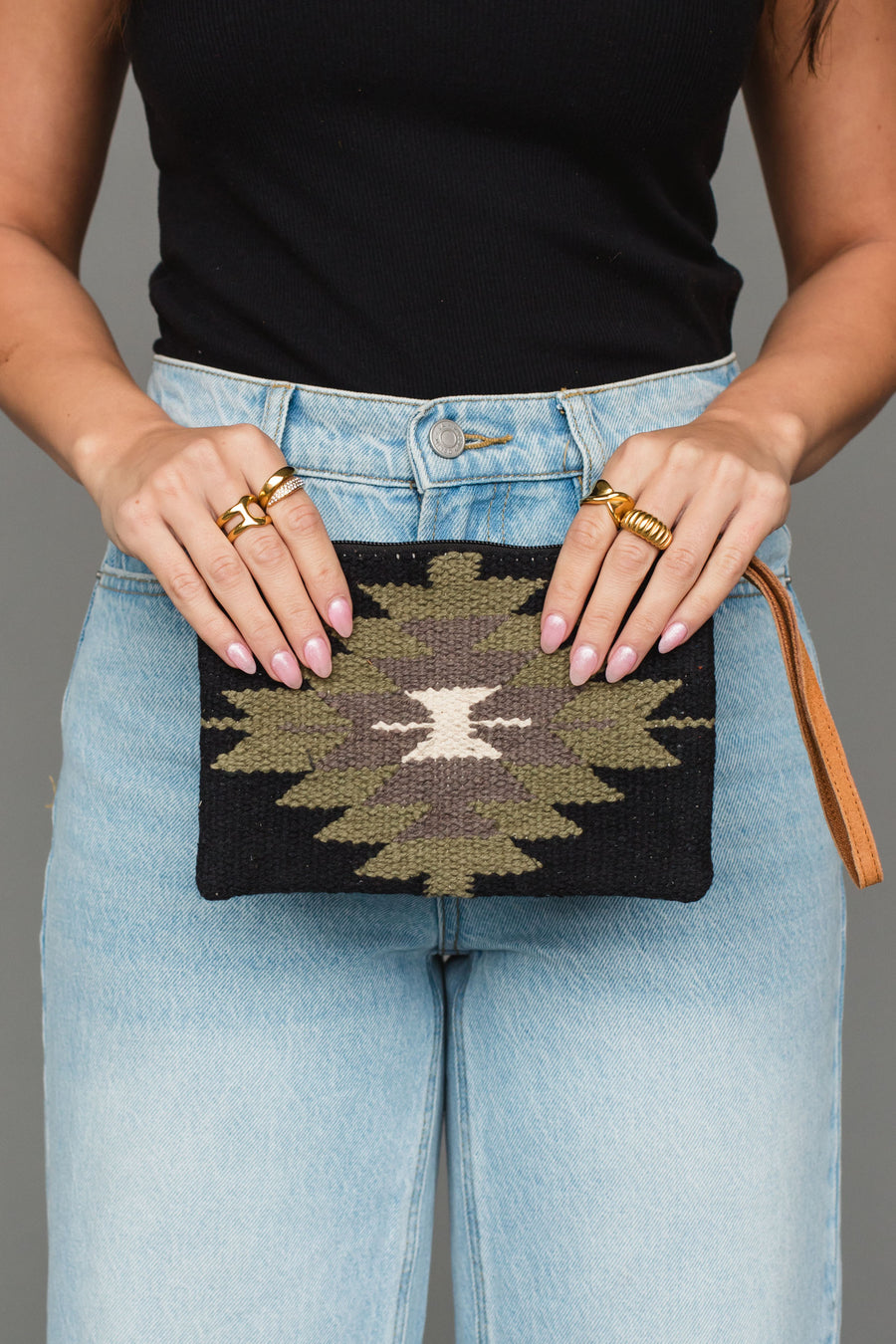 A person in a black sleeveless top and light blue jeans holds the Black, Olive, Gray & Cream Western Wristlet from Panache Apparel Collection, featuring a geometric pattern in earthy tones. They wear gold rings on both hands and have light pink nails.
