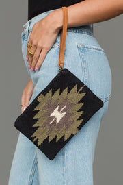 A person in light blue jeans holds the Panache Apparel Collection Black, Olive, Gray & Cream Western Wristlet, which features a geometric pattern and a tan wrist strap.