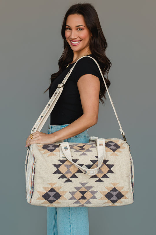 A woman in a black shirt and blue jeans smiles while holding the Panache Apparel Collection Khaki, Gray & Tan Western Duffel, featuring geometric patterns and shoulder straps, against a gray background.