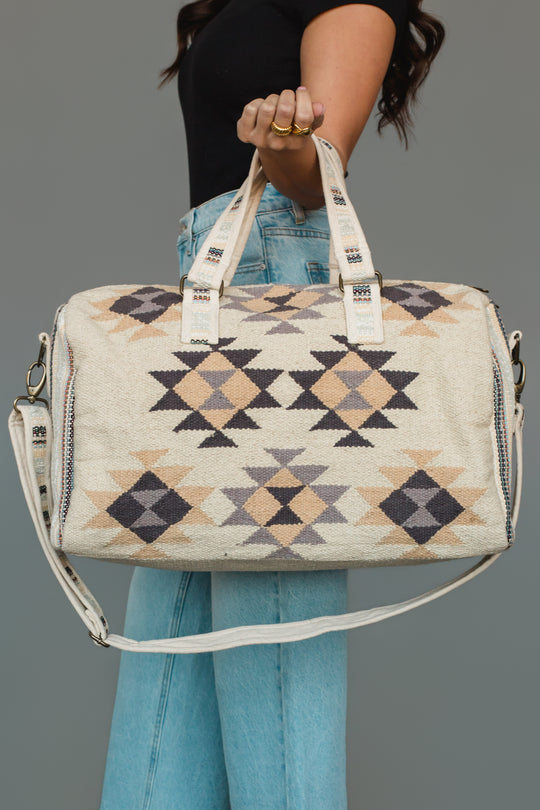 A woman in a black shirt and light blue jeans holds the Panache Apparel Collection Khaki, Gray & Tan Western Duffel, a large bag featuring geometric patterns in black, brown, and tan with both hand and shoulder straps.
