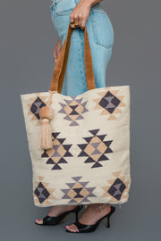 A person in jeans and heels holds the Panache Apparel Collection Khaki, Gray & Tan Western Tote, featuring geometric patterns and a tassel, against a gray background.