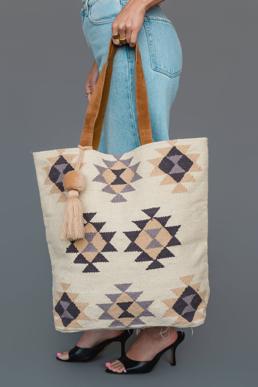 A person in jeans and heels holds the Panache Apparel Collection Khaki, Gray & Tan Western Tote, featuring geometric patterns and a tassel, against a gray background.