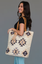 A woman smiles while holding the Khaki, Gray & Tan Western Tote from Panache Apparel Collection. The large tote showcases geometric patterns in beige, brown, and gray with brown straps and tassel accents. She pairs it with a black top and light blue jeans against a plain background.