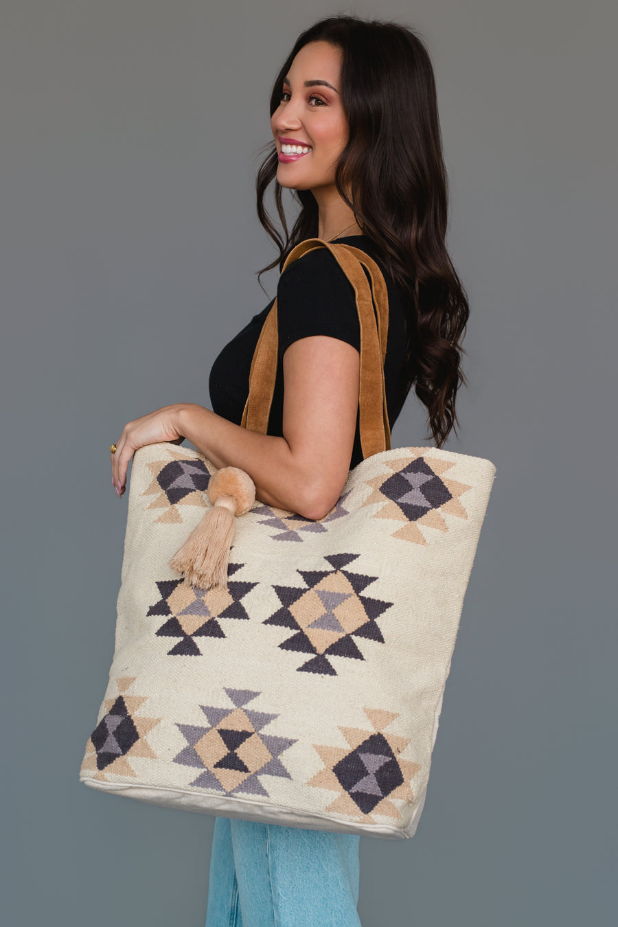 A woman smiles while holding the Khaki, Gray & Tan Western Tote from Panache Apparel Collection. The large tote showcases geometric patterns in beige, brown, and gray with brown straps and tassel accents. She pairs it with a black top and light blue jeans against a plain background.