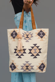 A person carrying the Panache Apparel Collection Khaki, Gray & Tan Western Tote, a woven tote bag featuring brown handles, geometric patterns in beige, tan, and black, and a tassel accent, paired with light blue jeans and a black top.