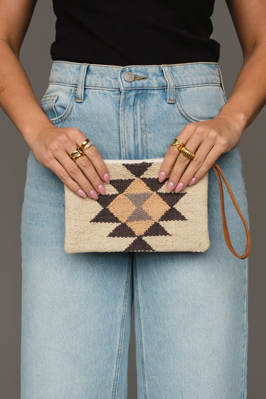 A person wearing light blue jeans and a black top holds the Panache Apparel Collection Khaki, Gray & Tan Western Wristlet, which features a woven geometric diamond pattern in black, beige, and tan. They have light pink nails and gold rings on both hands.