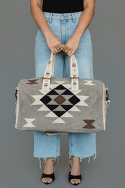 A person in light blue jeans and black sandals holds the Panache Apparel Collection Taupe, Cream, Brown & Black Western Duffel, standing against a plain gray background.