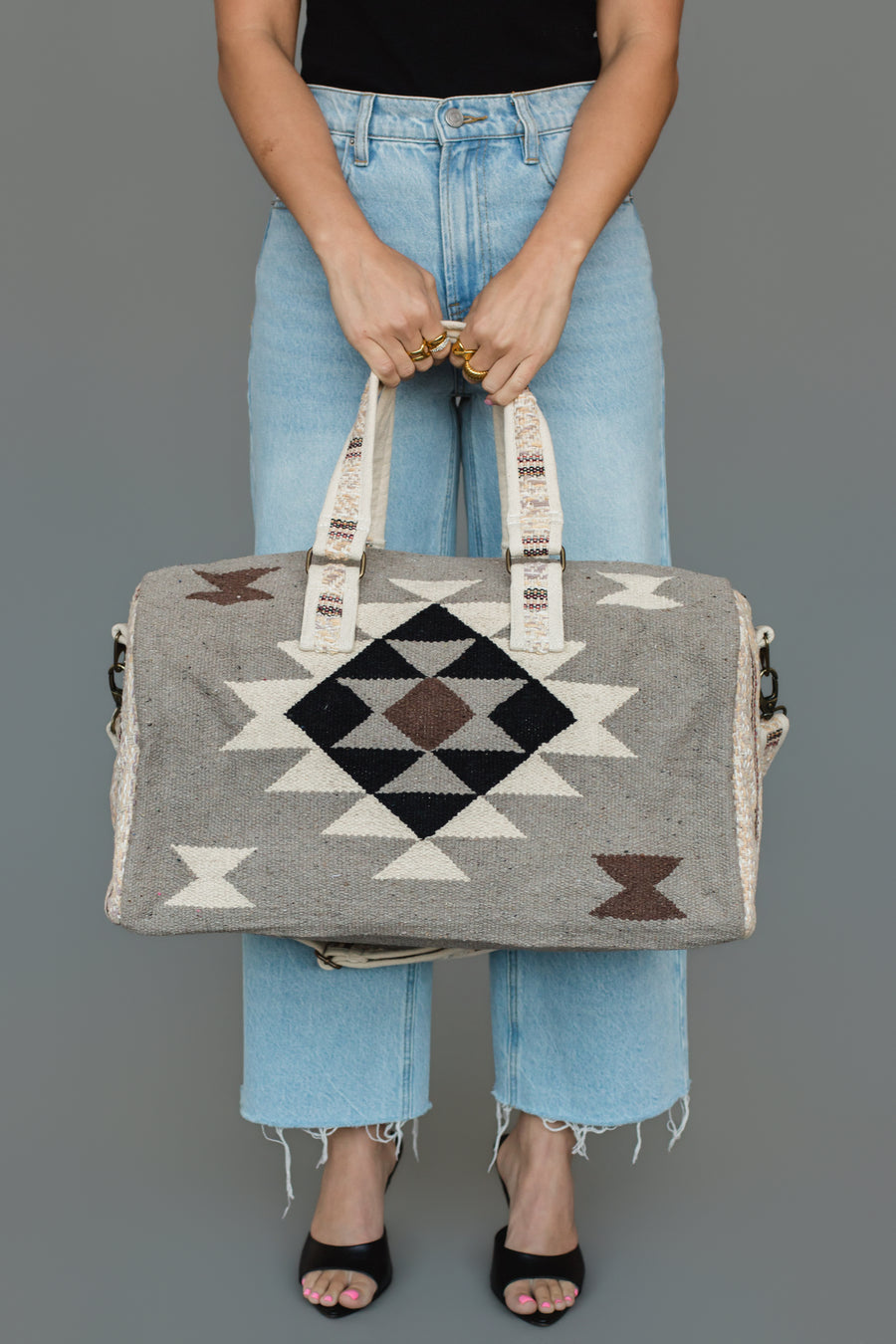 A person in light blue jeans and black sandals holds the Panache Apparel Collection Taupe, Cream, Brown & Black Western Duffel, standing against a plain gray background.