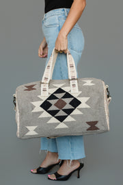 A person in blue jeans and black heels holds the Panache Apparel Collection Taupe, Cream, Brown & Black Western Duffel, featuring geometric patterns and cream-colored handles, against a plain gray background.