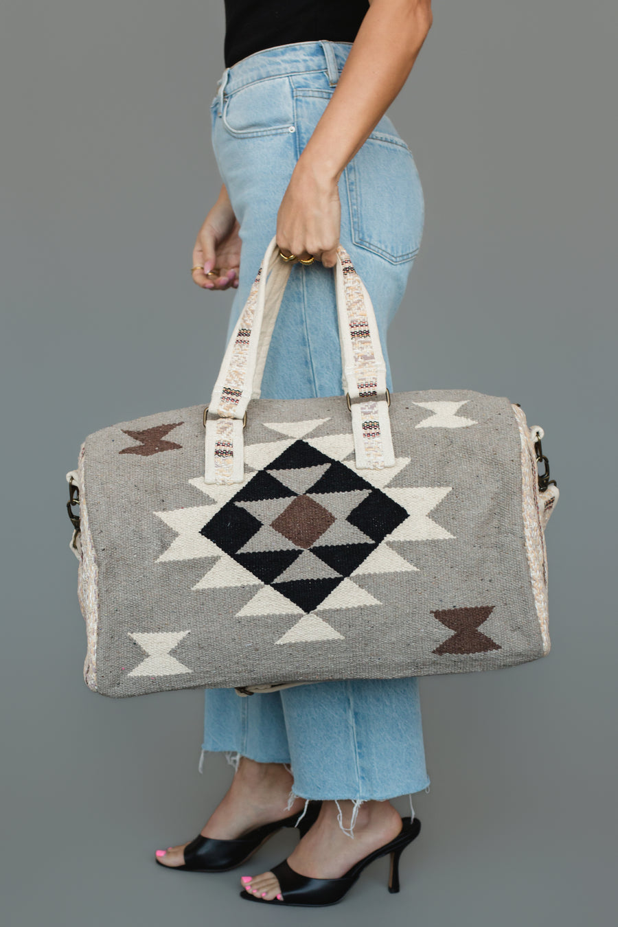 A person in blue jeans and black heels holds the Panache Apparel Collection Taupe, Cream, Brown & Black Western Duffel, featuring geometric patterns and cream-colored handles, against a plain gray background.