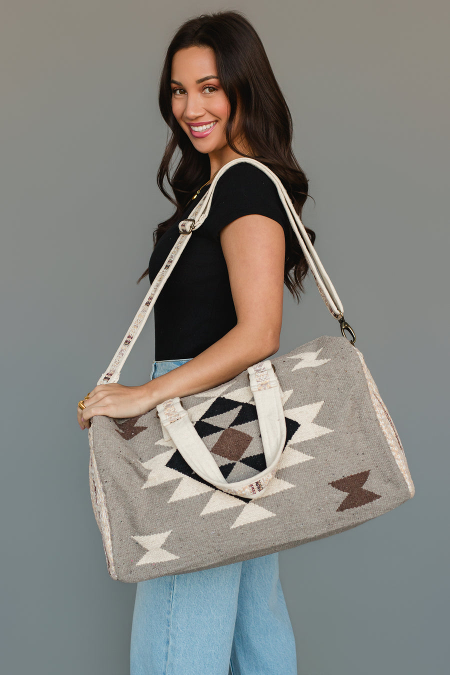 A woman with long dark hair, dressed in a black top and light blue jeans, smiles while holding the Panache Apparel Collection Taupe, Cream, Brown & Black Western Duffel over her shoulder against a plain gray background.