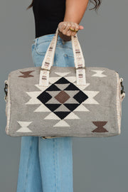 A person in blue jeans and a black top holds the Panache Apparel Collection’s Taupe, Cream, Brown & Black Western Duffel, featuring geometric patterns and cream handles with text.