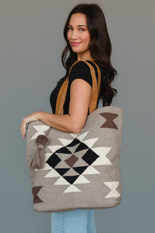 A woman with long dark hair in a black top and jeans holds the Panache Apparel Collection’s Taupe, Cream, Brown & Black Western Tote, featuring geometric patterns and a tassel, while standing against a plain gray background.