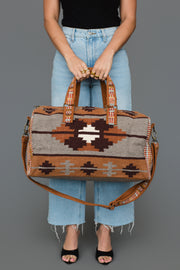 A woman wearing a black top and light blue jeans holds the Rust, Gray & Brown Western Duffel by Panache Apparel Collection, a large bag featuring geometric patterns in brown and beige with a leather strap.