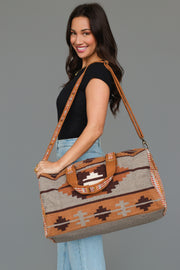 A smiling woman with long brown hair, dressed in a black top and jeans, holds the Panache Apparel Collection Rust, Gray & Brown Western Duffel, a large patterned bag with a shoulder strap, against a plain gray background.
