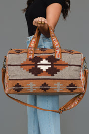 A person holds the Panache Apparel Collection Rust, Gray & Brown Western Duffel, featuring bold geometric southwestern patterns in shades of brown, rust, gray, and white. They wear light jeans and a black shirt, standing against a plain gray background.