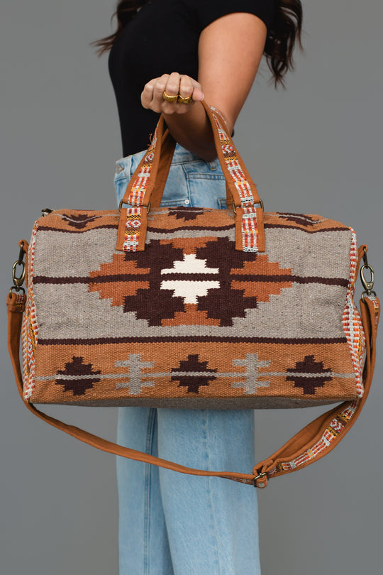 A person holds the Panache Apparel Collection Rust, Gray & Brown Western Duffel, featuring bold geometric southwestern patterns in shades of brown, rust, gray, and white. They wear light jeans and a black shirt, standing against a plain gray background.
