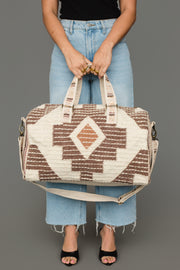 A person in light blue jeans and black sandals holds the Panache Apparel Collection Cream, Brown & Rust Western Duffel, featuring geometric patterns and a shoulder strap, against a gray background.