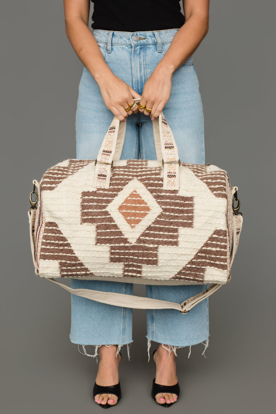A person in light blue jeans and black sandals holds the Panache Apparel Collection Cream, Brown & Rust Western Duffel, featuring geometric patterns and a shoulder strap, against a gray background.