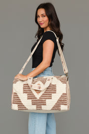 A woman with long dark hair wears a black top and light blue jeans, carrying the Panache Apparel Collection’s Cream, Brown & Rust Western Duffel, a large patterned bag with shoulder and hand straps, while standing against a plain gray background.