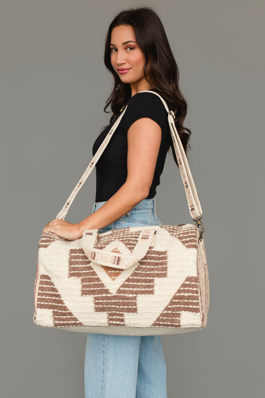 A woman with long dark hair wears a black top and light blue jeans, carrying the Panache Apparel Collection’s Cream, Brown & Rust Western Duffel, a large patterned bag with shoulder and hand straps, while standing against a plain gray background.