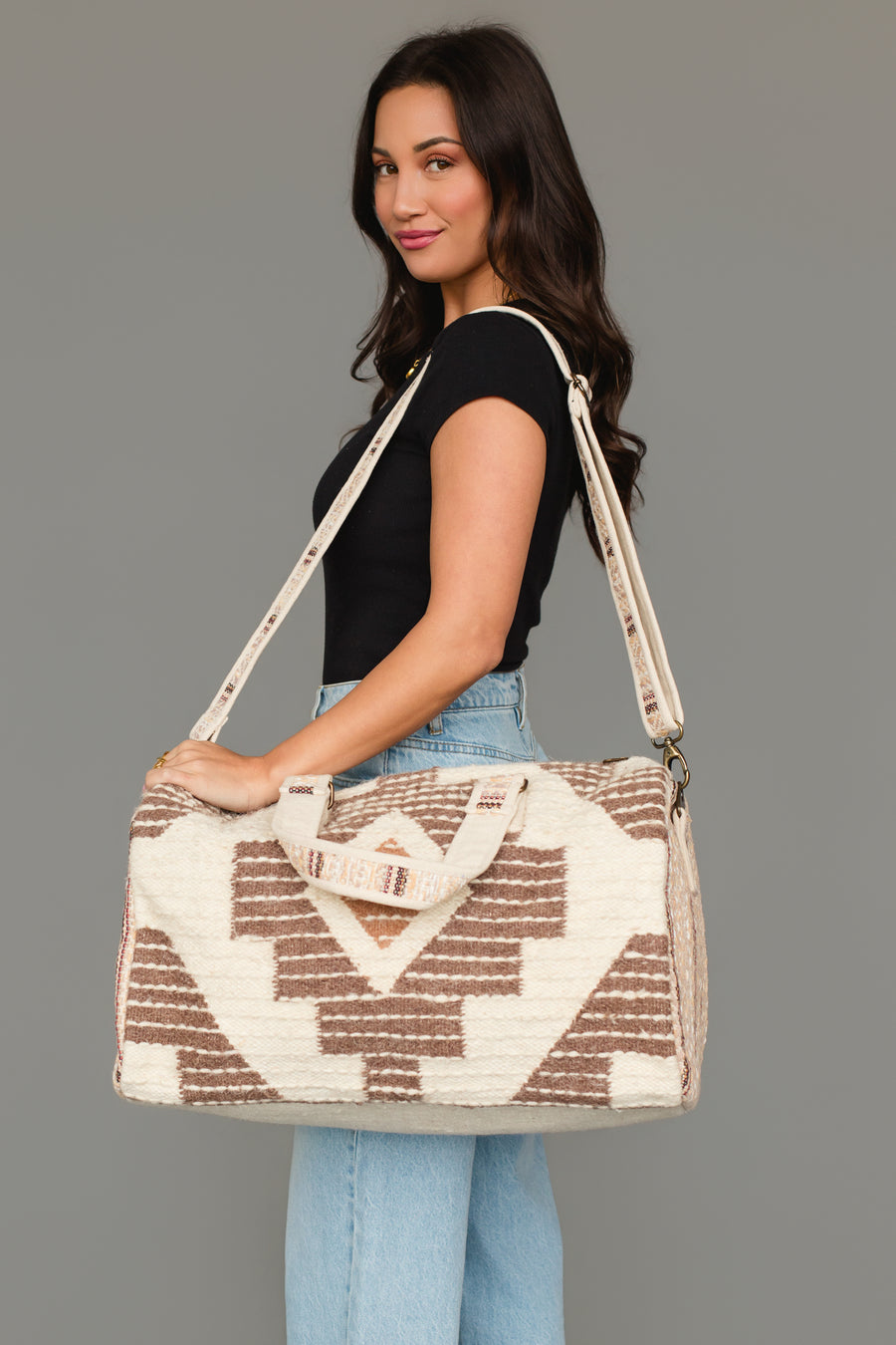 A woman with long dark hair wears a black top and light blue jeans, carrying the Panache Apparel Collection’s Cream, Brown & Rust Western Duffel, a large patterned bag with shoulder and hand straps, while standing against a plain gray background.