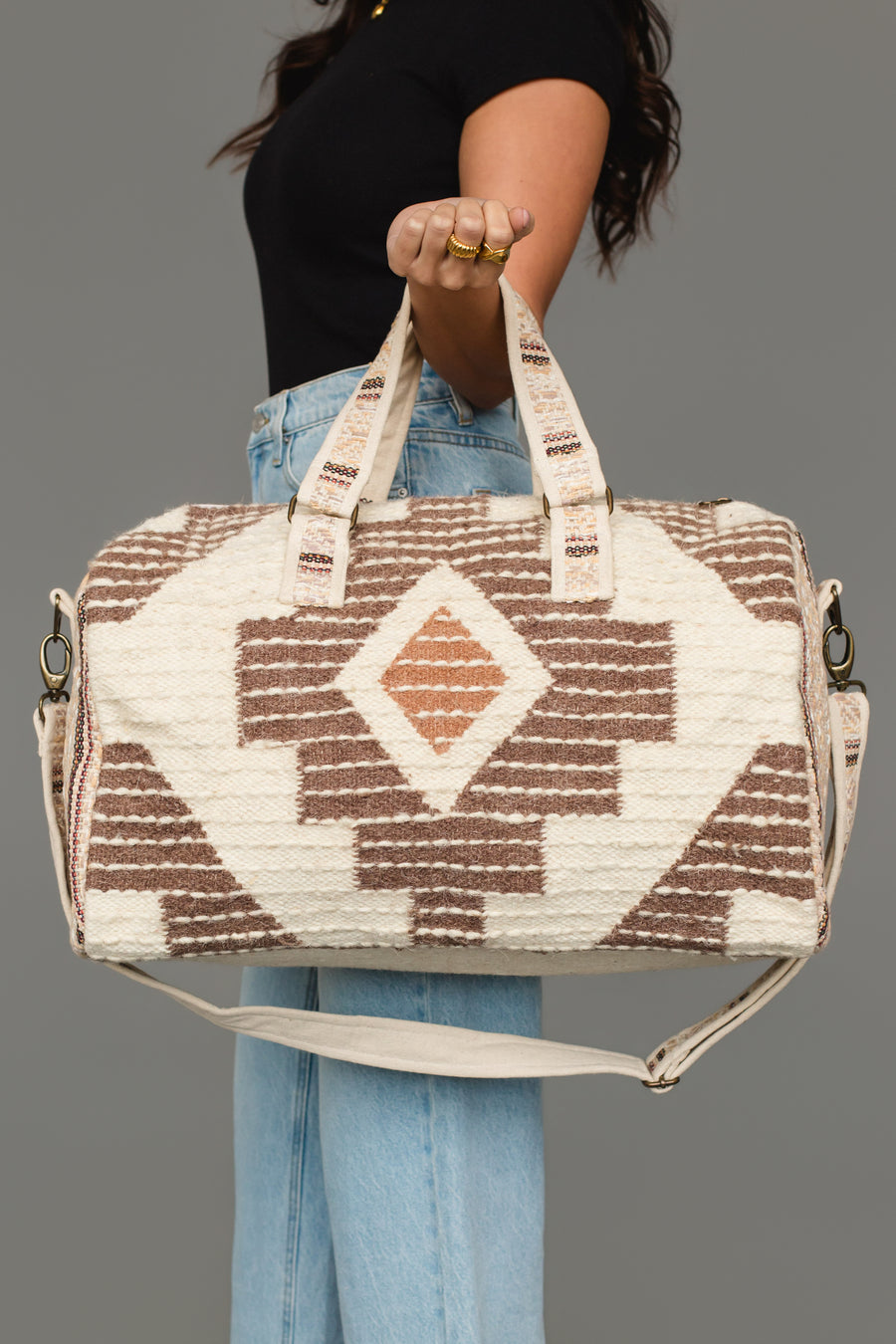 A person in a black top and blue jeans holds the Cream, Brown & Rust Western Duffel by Panache Apparel Collection, featuring geometric patterns and two handles, against a plain gray background.