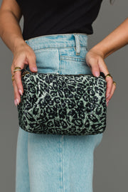 A person dressed in light blue jeans and a black top holds the Panache Apparel Collection Green & Black Leopard Travel Pouch with both hands against a plain gray background.