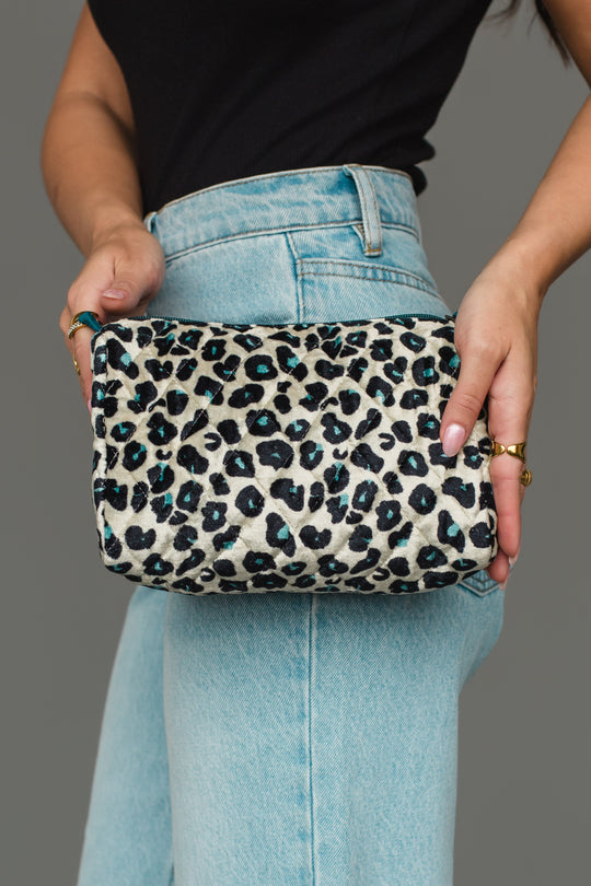 A person wearing light blue jeans and a black top holds the Panache Apparel Collection Black, Cream & Blue Leopard Travel Pouch with both hands in front of a gray background.