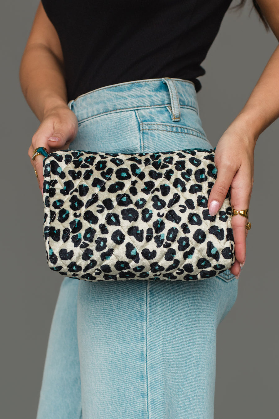 A person wearing light blue jeans and a black top holds the Panache Apparel Collection Black, Cream & Blue Leopard Travel Pouch with both hands in front of a gray background.