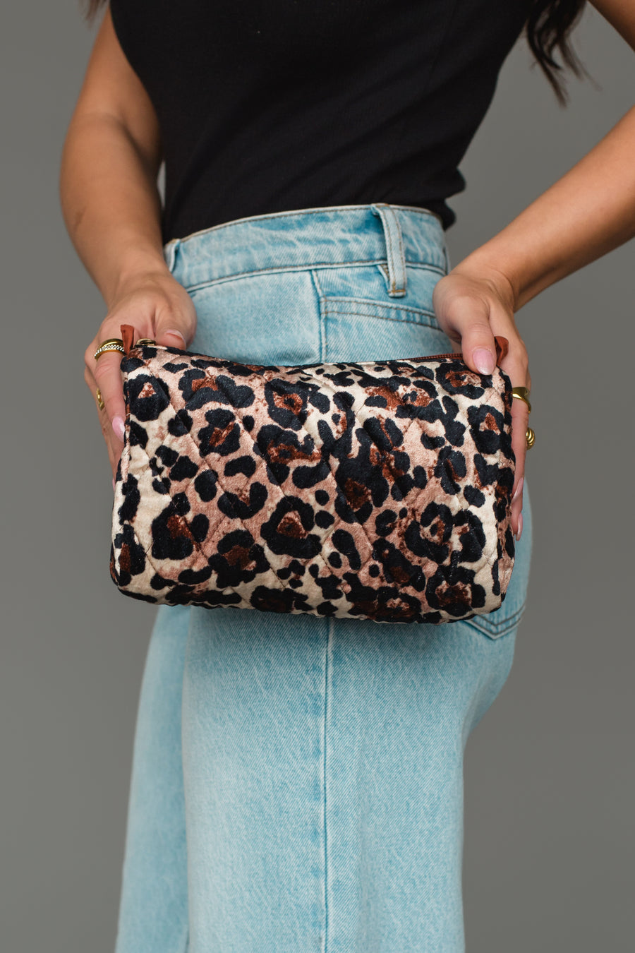 A person in a black top and light blue jeans holds the Panache Apparel Collection Brown & Cream Leopard Travel Pouch with both hands against a plain gray background.
