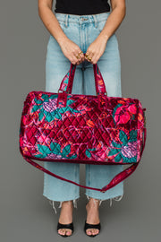 A person holds the Panache Apparel Collection Burgundy & Multicolored Floral Duffel, featuring red, pink, teal, and purple tones. They wear light blue jeans, a black top, and black open-toe shoes against a plain gray background.
