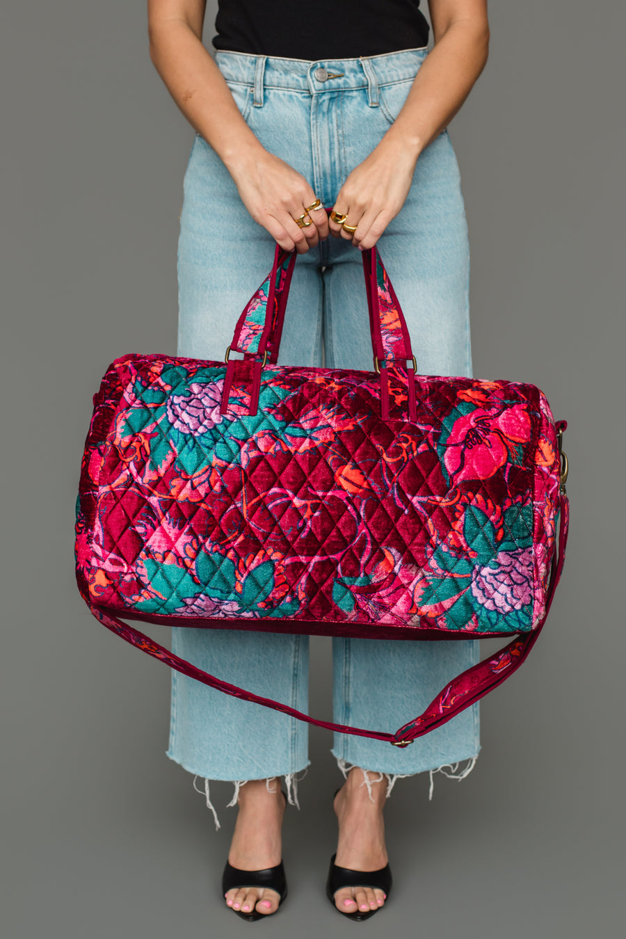 A person holds the Panache Apparel Collection Burgundy & Multicolored Floral Duffel, featuring red, pink, teal, and purple tones. They wear light blue jeans, a black top, and black open-toe shoes against a plain gray background.